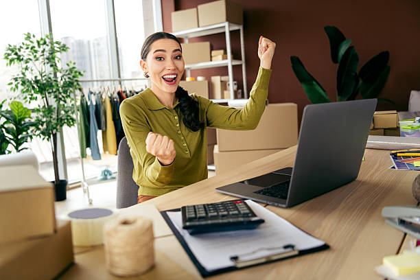 زنی جوان که در دفتر کار خود، موفقیت کسب و کار آنلاینش را جشن میگیرد زنی جوان که در دفتر کار خود، موفقیت کسب و کار آنلاینش را جشن میگیرد